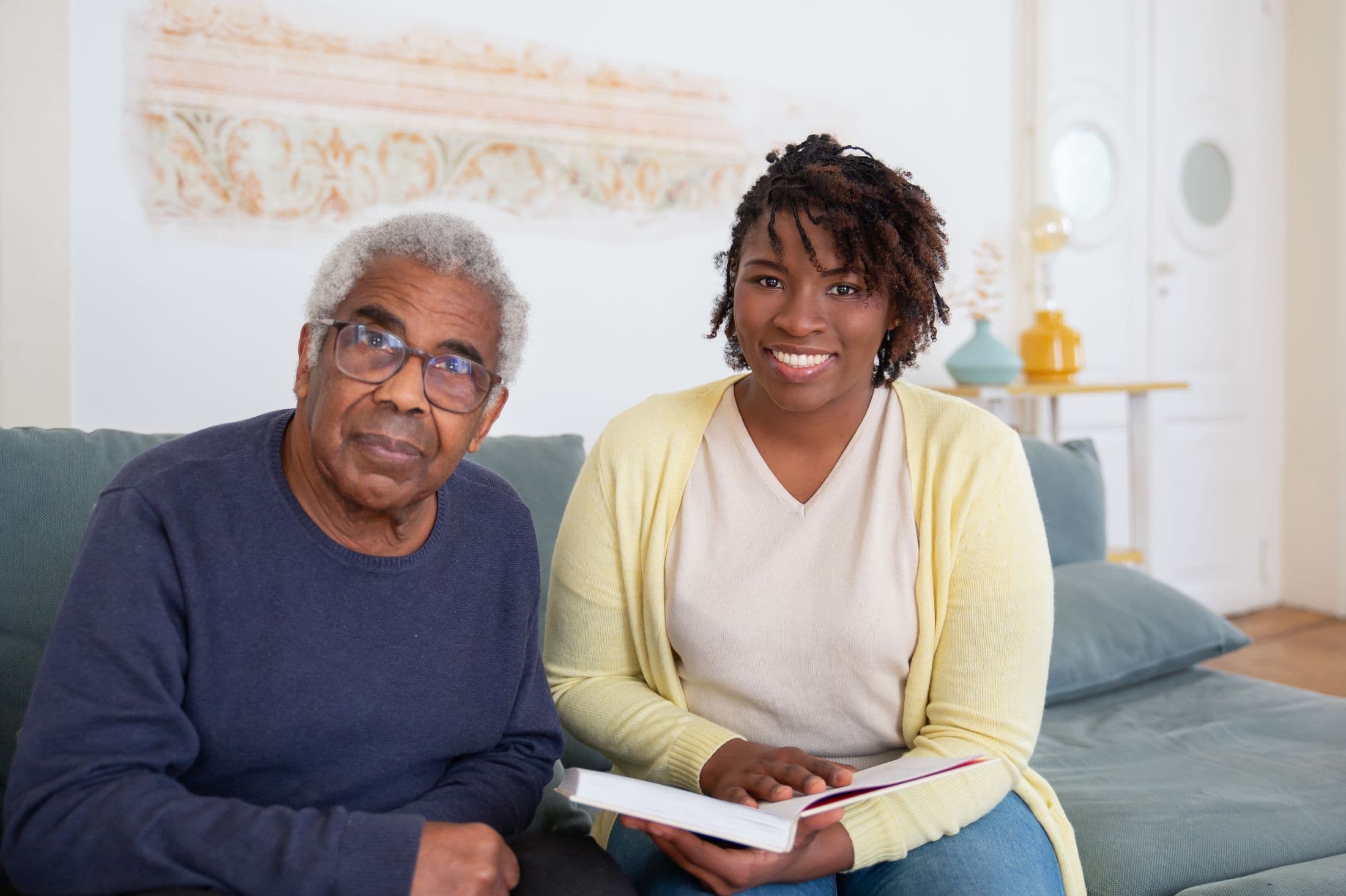 A caregiver and an older man smiling together on the sofa at home